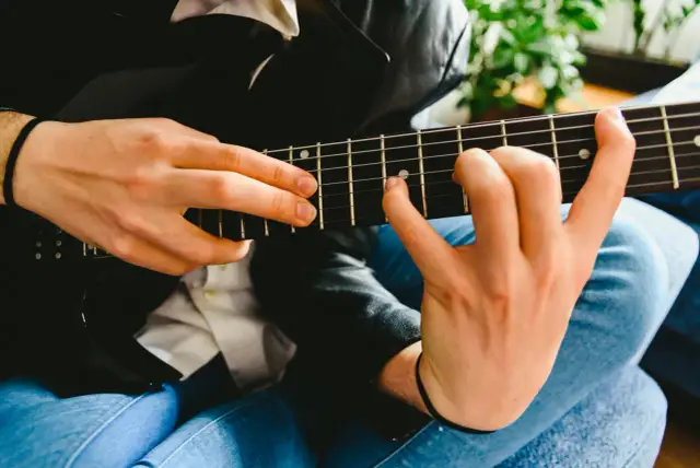 Dłonie grające na czarnej gitarze elektrycznej, z widocznymi progami i strunami.