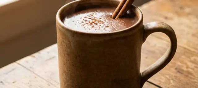 Eine dampfende Tasse vegane heiße Schokolade mit Zimtstange und Kakaopulver auf einem Holztisch.