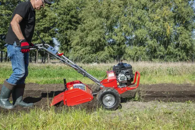 Mężczyzna w rękawiczkach i gumowcach obsługuje czerwoną glebogryzarkę na polu.