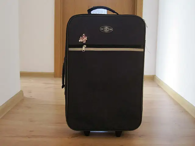 Czarna walizka na kółkach z ozdobnym przypinką, stojąca na drewnianej podłodze.