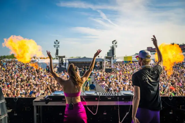DJ-ka i DJ na scenie, płomienie buchają, tłum szaleje na festiwalu muzyki elektronicznej.