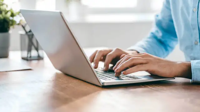 Person tippt auf einem silbernen Laptop auf einem Holztisch.