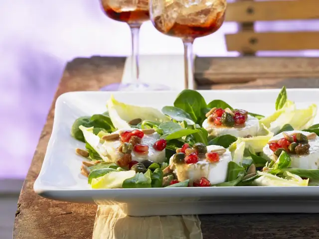 Ein frischer Salat mit Ziegenkäse, garniert mit Granatapfelkernen und Pinienkernen, serviert auf einem weißen Teller.