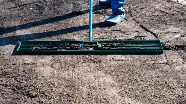 Osoba w niebieskich spodniach i gumowcach wyrównuje ziemię pod trawnik za pomocą zielonej równiarki.