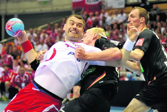 Gracz w białej koszulce z piłką ręczną próbuje wykonać rzut karny, podczas gdy przeciwnik próbuje go zatrzymać.