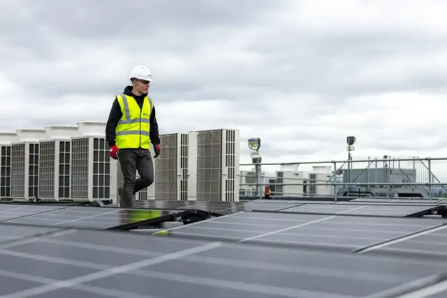 Pracownik w kamizelce odblaskowej przechodzi obok rzędu klimatyzatorów na dachu, gdzie instalowane są panele słoneczne. sofar solar polska.