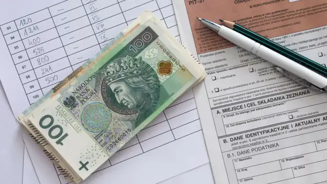 Stos banknotów 100 zł leży na formularzu PIT. Obok arkusz z danymi i ołówki.