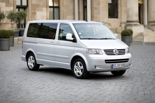 Srebrny Volkswagen T5 na brukowanym placu przed budynkiem z kolumnami.