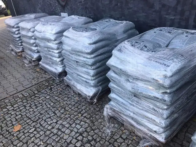 Stosy worków z asfaltem na paletach, owinięte folią, na kamiennym podłożu.