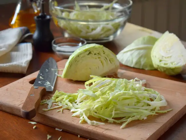 Frisch geschnittener Weißkohl liegt auf einem Holzbrett, bereit für ein Sauerkraut Rezept. Daneben ein Messer und Schüssel mit Kohl.