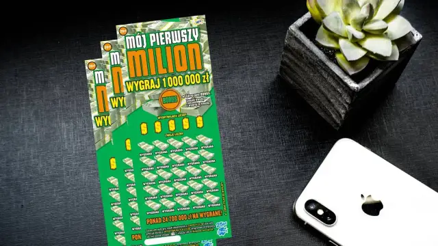Zielona zdrapka "Mój pierwszy milion" z napisem "Wygraj 1 000 000 zł" leży obok białego iPhone'a i doniczki z sukulentem.