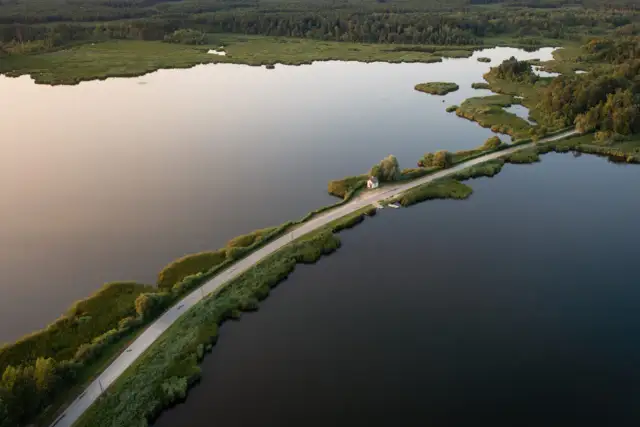 Droga przecinająca jezioro, otoczona zielenią i lasem. W oddali mały domek, jakby cyców na wodzie.