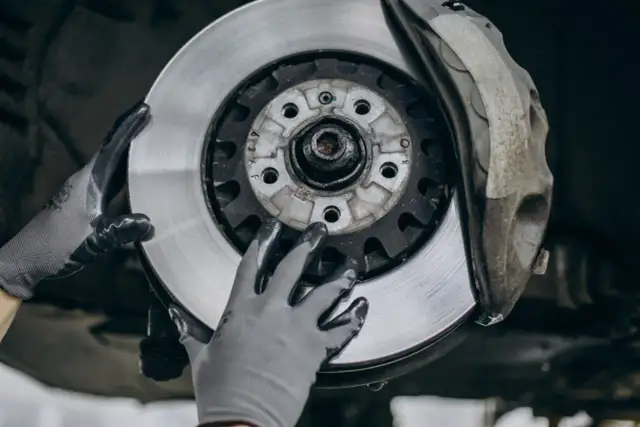 Mechanik w rękawiczkach wymienia zużyte tarcze hamulcowe. Widać nowy, lśniący element obok starego zacisku.