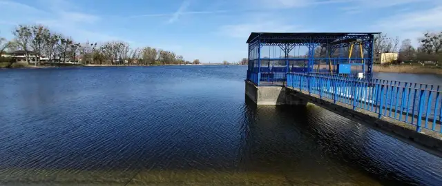 Spokojne jezioro średzkie z błękitną budowlą i pomostem. W tle drzewa i zabudowania.