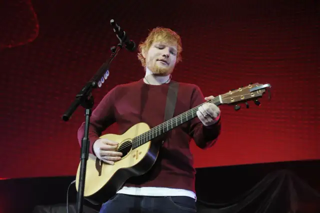 Ed Sheeran gra na gitarze akustycznej na tle czerwonej sceny.