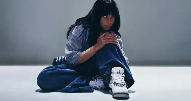 Billie Eilish w luźnych dżinsach i koszuli, zamyślona, w butach Jordan. Tekst "What was I made for?" pasuje do jej nastroju.