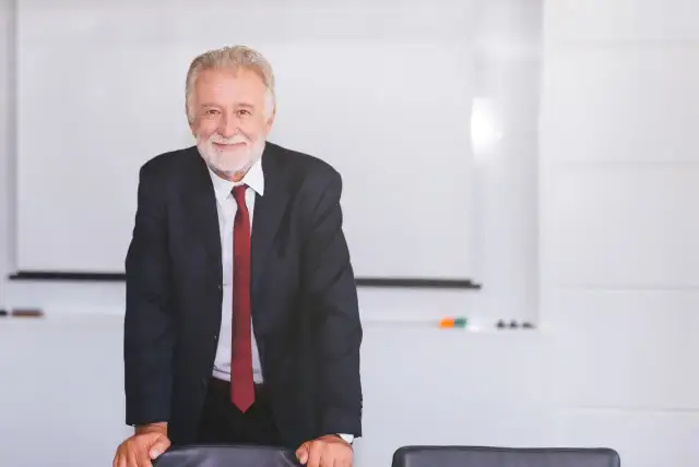 Uśmiechnięty starszy mężczyzna w garniturze stoi przy stole w sali konferencyjnej.