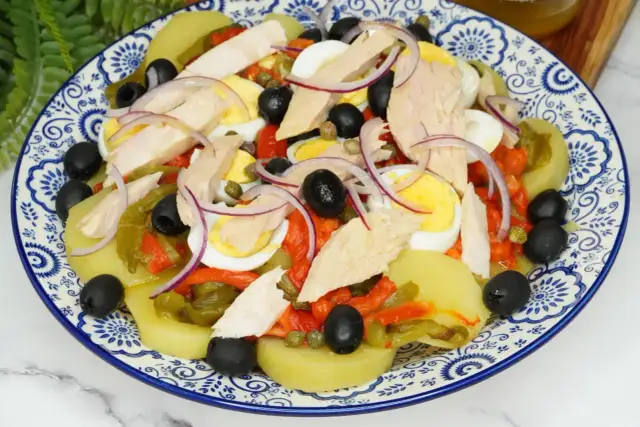 Deliciosa ensalada de patata con atún, huevo duro, pimientos asados, aceitunas negras y cebolla morada.