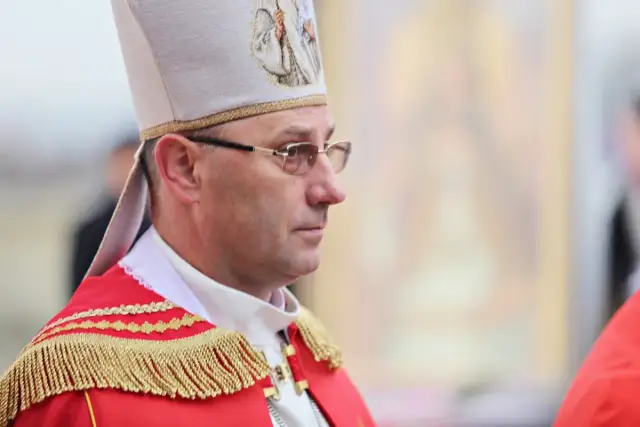Biskup w stroju pontyfikalnym, z mitrą na głowie, w okularach. W tle widać fragment obrazu.