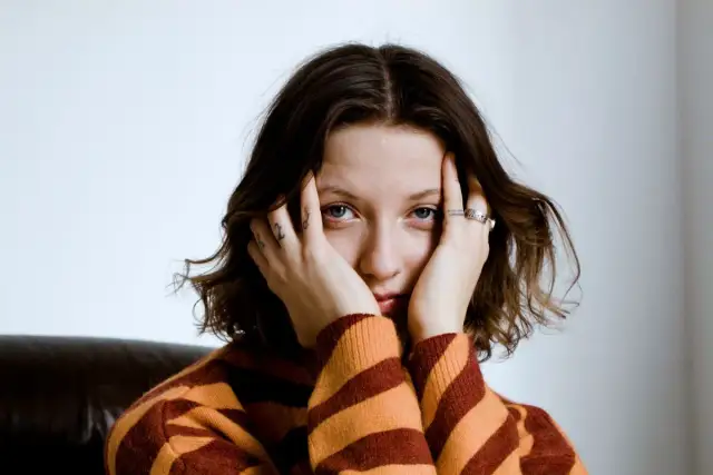 Justyna Święs Piernikowski w swetrze w pasy, z dłońmi przy twarzy, patrzy w obiektyw.