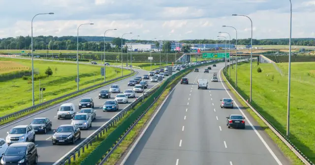 Autostrada z ruchem samochodowym w słoneczny dzień, z zielonymi poboczami i drzewami w tle.