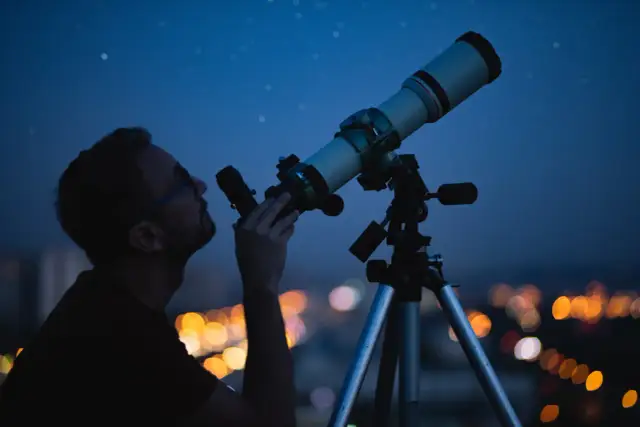 Mężczyzna obserwuje nocne niebo przez teleskop na tle rozświetlonego miasta.