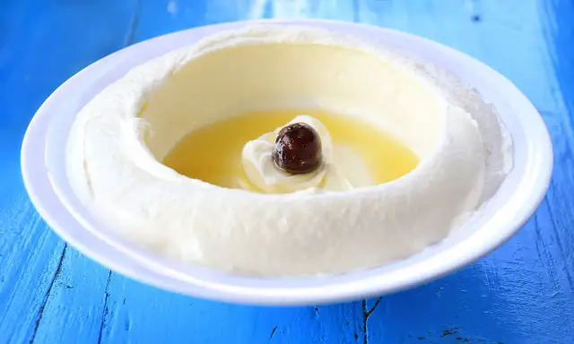 Un plato blanco con labneh que es un queso cremoso, con aceite de oliva y una aceituna negra en el centro, sobre una mesa azul.