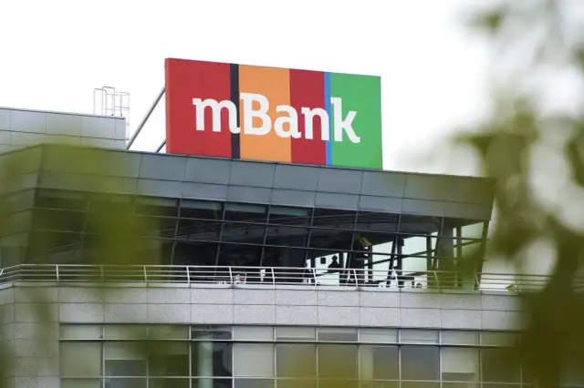 Kolorowy szyld z napisem mBank na dachu nowoczesnego budynku. To siedziba banku, do kogo należy mbank, jest jasne dla każdego.