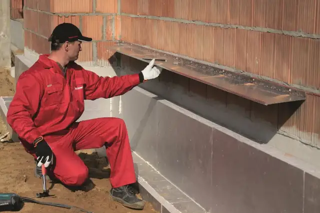 Pracownik w czerwonym kombinezonie montuje okapnik na ścianie zewnętrznej, przygotowując ją do montażu wełny mineralnej.