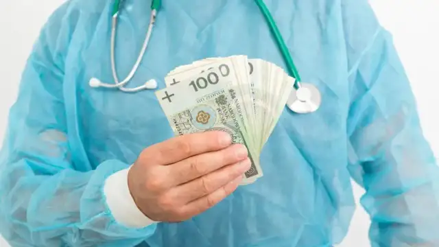 Lekarz rezydent w niebieskim fartuchu trzyma w dłoni plik banknotów 100 zł. Pytanie brzmi: ile zarabia lekarz rezydent?