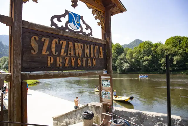 Drewniany szyld "Szczawnica Przystań" nad rzeką z tratwami i górami w tle.