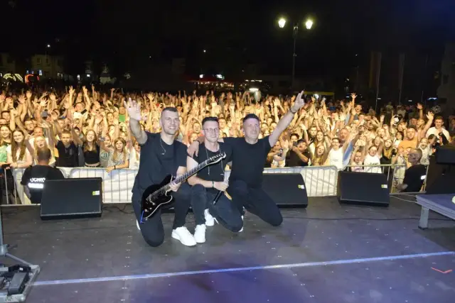 Trzech muzyków na scenie, z gitarą i mikrofonem, pozuje przed rozentuzjazmowanym tłumem. To z pewnością udane imieniny Jana!