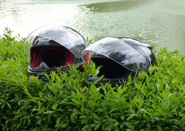 Dwa kaski motocyklowe w zielonych liściach, z wodą w tle.