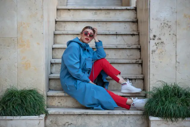 Kobieta w niebieskim płaszczu i czerwonych spodniach, w białych sneakersach, prezentuje sportową elegancję na kamiennych schodach.