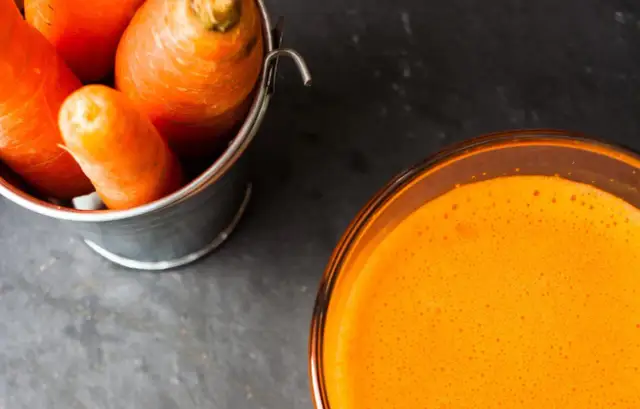 Frische Karotten in einem Eimer neben einem Glas leuchtend orangem Karottensaft. Die Wirkung von Karottensaft ist bekannt für seine Vitamine.
