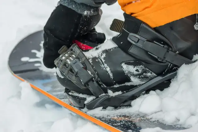 Rękawica zapina but snowboardowy na desce, przygotowując się do wygrzewania butów snowboardowych w śniegu.