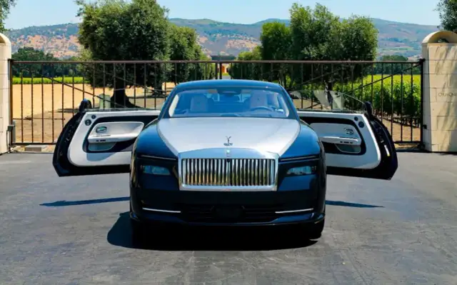 Elegancki Rolls-Royce Spectre z otwartymi drzwiami, zaparkowany przed bramą posiadłości z widokiem na wzgórza.