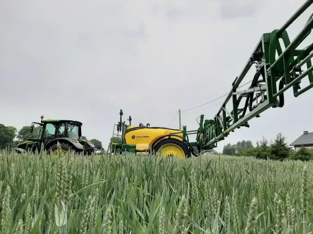 Traktor John Deere z opryskiwaczem w polu pszenicy, gotowy do zabiegu z użyciem środków ochrony roślin, w tym glifosatu.