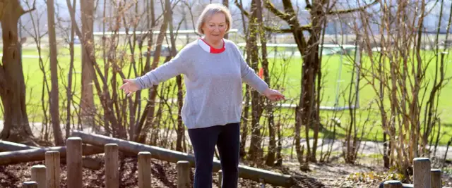 Ältere Frau balanciert auf Holzstämmen im Freien.