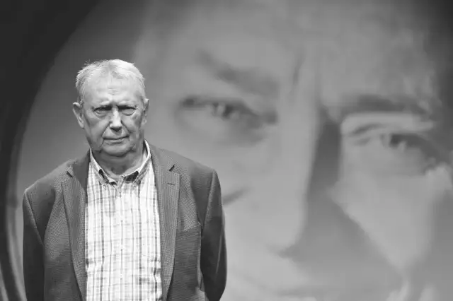 Wojciech Młynarski, jakby z tekstu "Jesteśmy na wczasach", w marynarce i kratowanej koszuli, na tle swojego portretu.