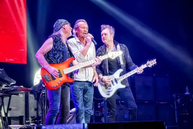 Trzech muzyków na scenie w głębokim fioletowym świetle Atlas Areny. Jeden śpiewa, dwaj grają na gitarach.