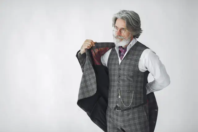 Elegancki mężczyzna z siwą brodą i wąsami, ubrany w kraciasty garnitur, prezentuje się jak prawdziwy arbiter elegantiarum.
