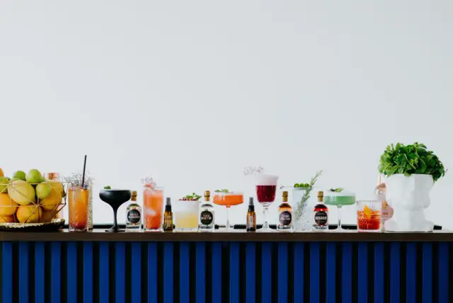 Rząd kolorowych drinków i butelek z alkoholami na barze, obok koszyka z owocami i białej rzeźby głowy.