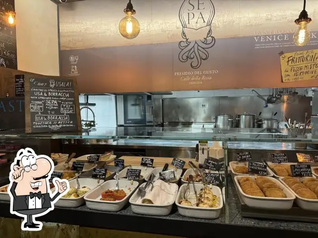 A display case filled with small dishes of food, with a chalkboard menu and a restaurant logo in the background.
