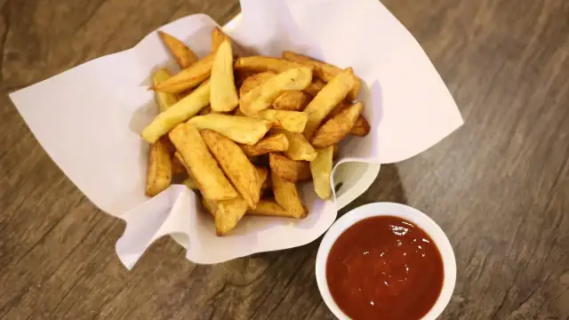 Chrupiące frytki stekowe w papierowym rożku obok miseczki z ketchupem na drewnianym tle.