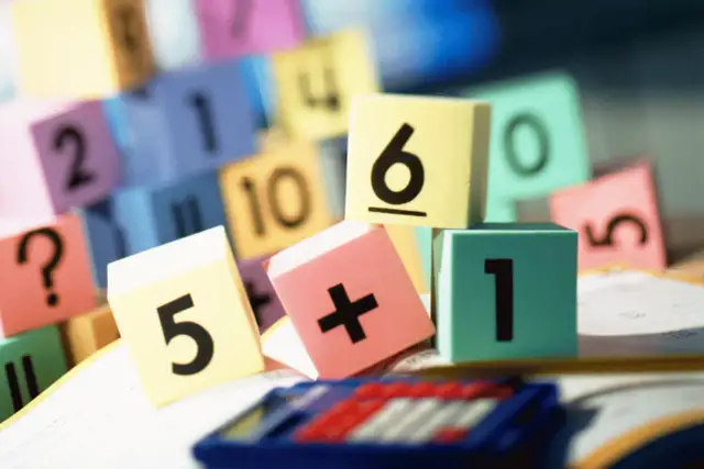 Kolorowe klocki z cyframi i znakami matematycznymi, leżące na kartce papieru obok kalkulatora.