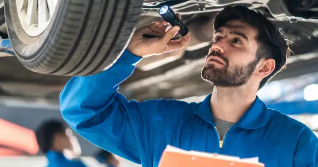 Mechanik w niebieskim kombinezonie oświetla latarką podwozie samochodu, wykonując **serwisowanie samochodu**.