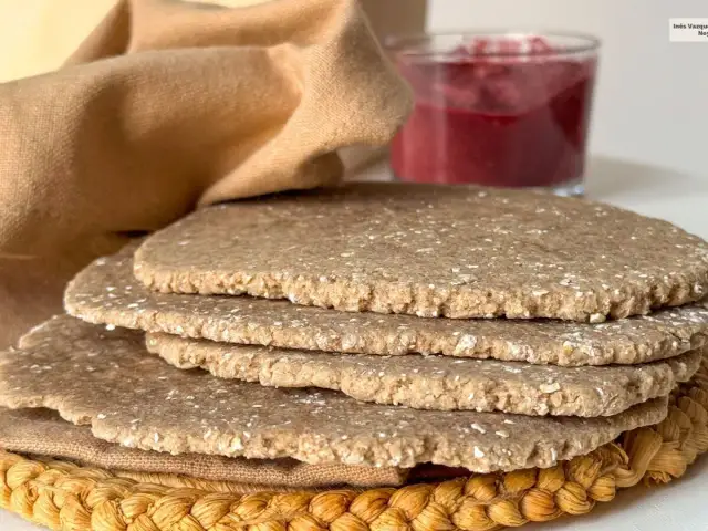 Una pila de pan sin levadura, rústico y con trozos de avena, junto a un vaso de mermelada de frutos rojos y una tela marrón.