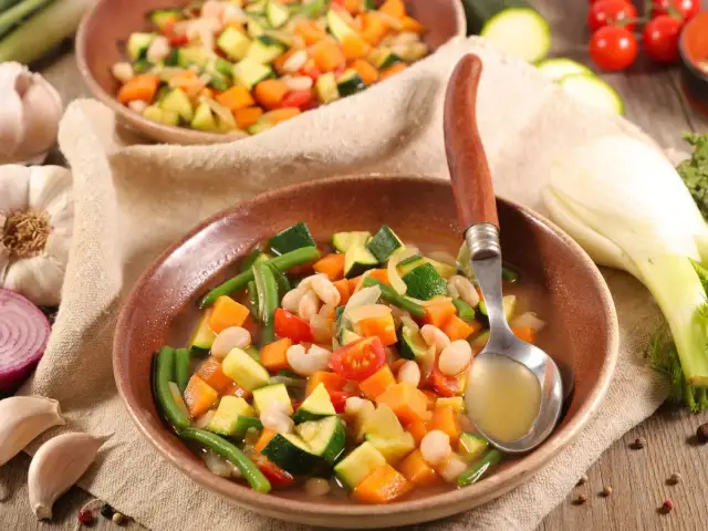 Un plato humeante de menestra de verduras, con judías verdes, zanahoria, calabacín y alubias blancas, servido con una cuchara.