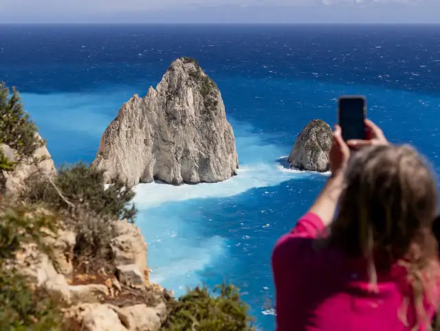 Widok na turkusowe morze i skaliste wysepki. Kobieta robi zdjęcie telefonem. Zastanawiasz się, ile się leci na Zakynthos?
